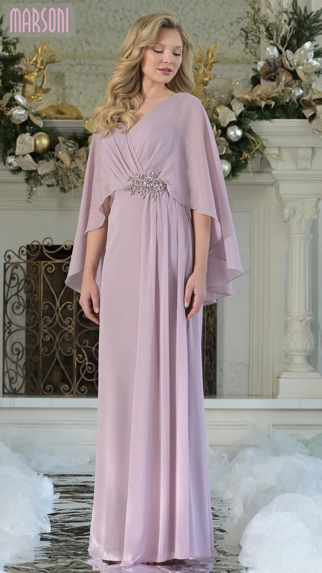 A woman in a floor-length, dusty rose Marsoni MV1361 formal dress with cape sleeves and a brooch at the waist stands in an elegant room adorned with white and gold floral arrangements.